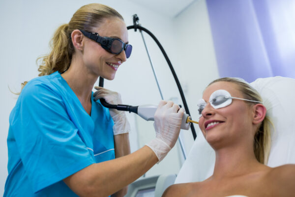 Kosmetikerin führt CO₂-Laserbehandlung im Gesicht einer lächelnden Patientin durch